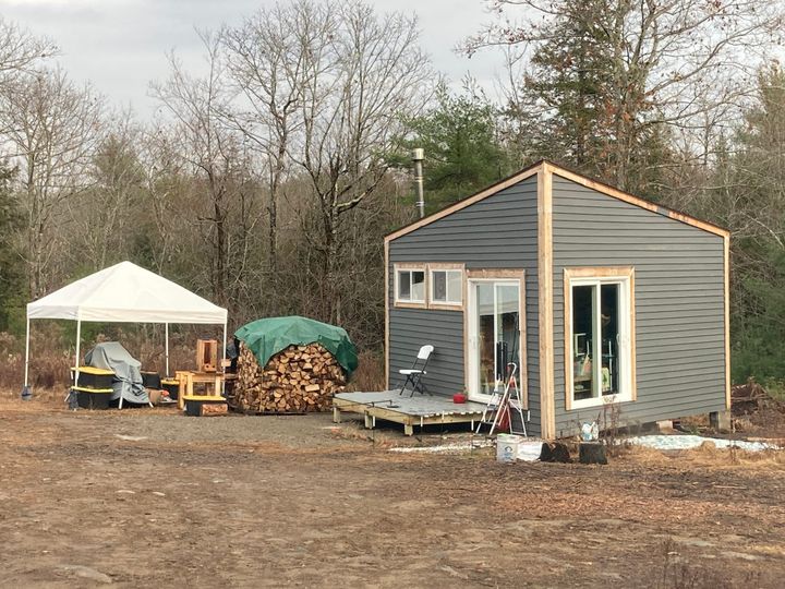 A homestead with a grey-sided house, unfinished deck, a round rik of firewood, and a canopy over tools.