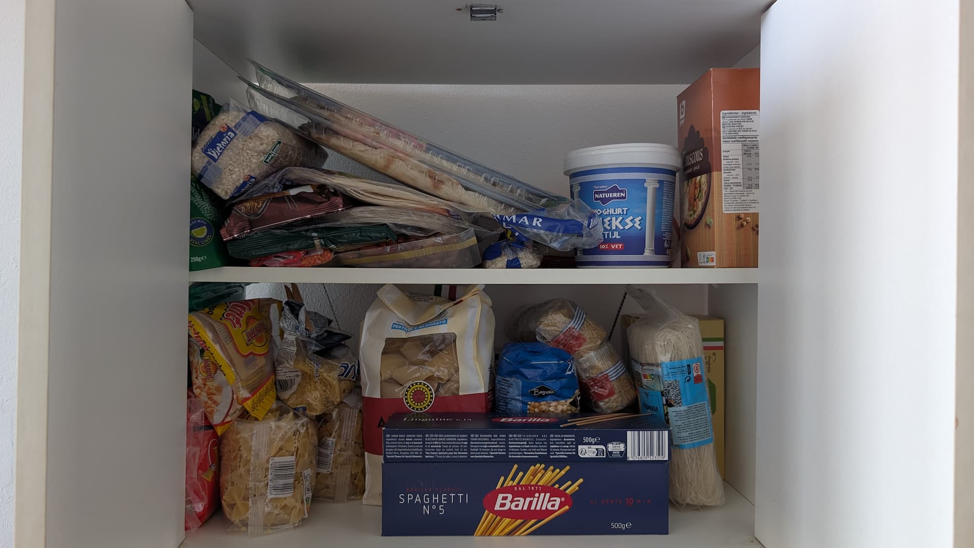 Shelving unit with bulk bought items, including packs of pasta, rice, wraps, pizza bases, noodles, couscous, and more.