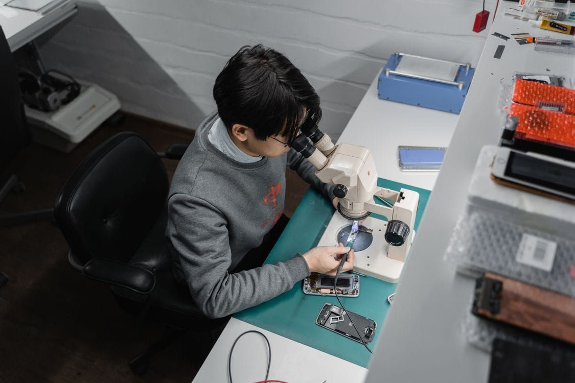 Technician using a microscope while repairing an opened smartphone at a workbench.