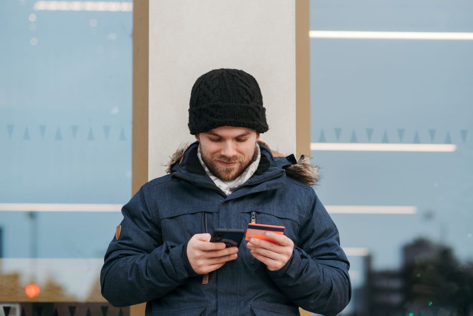 A man standing outside uses a phone while holding a payment card - click-to-cancel laws in 2026