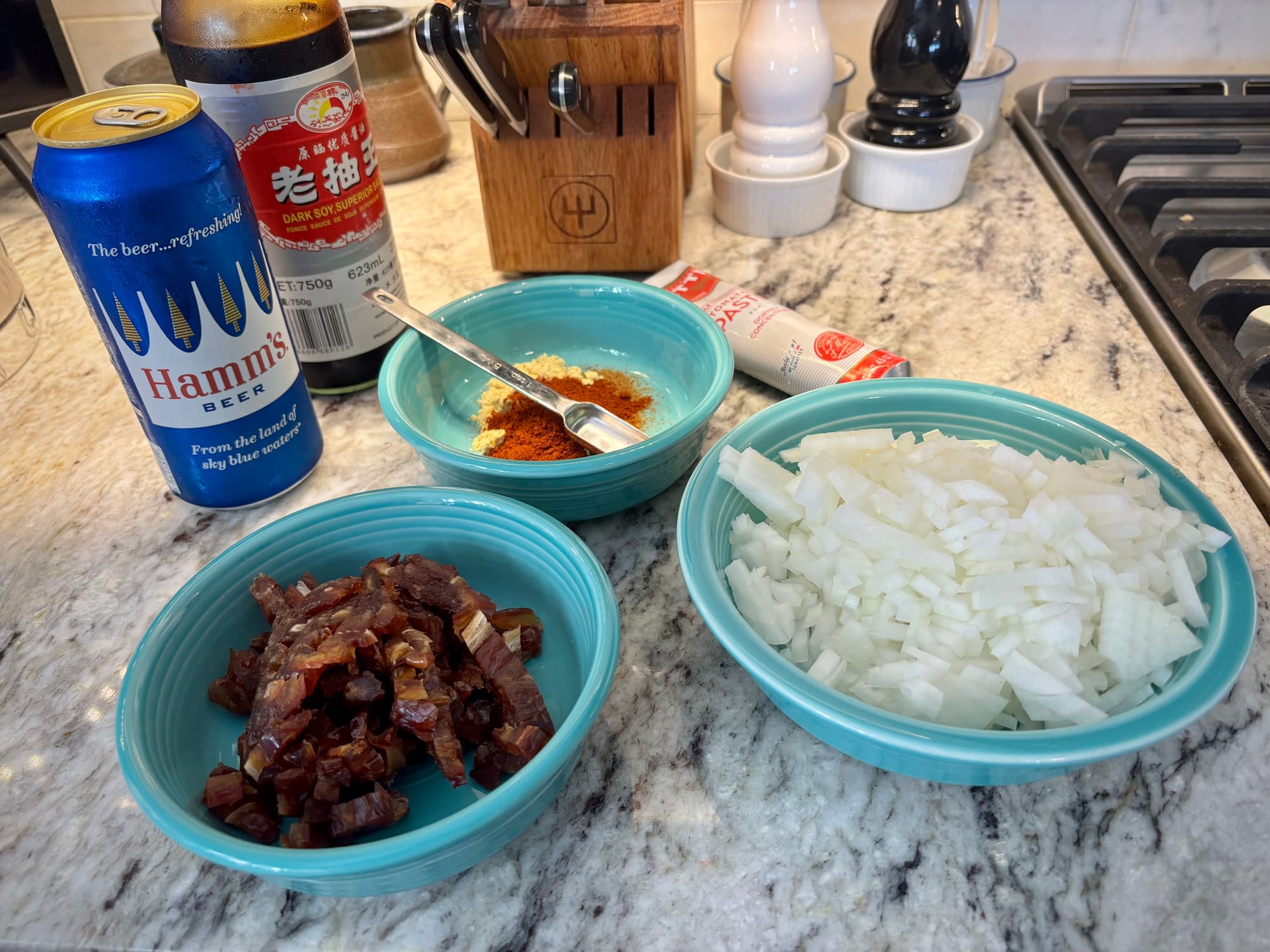 A can of beer, bottle of dark soy sauce, and a tube of tomato paste, with three blue ceramic bowls filled with spices, chopped dates and diced onions