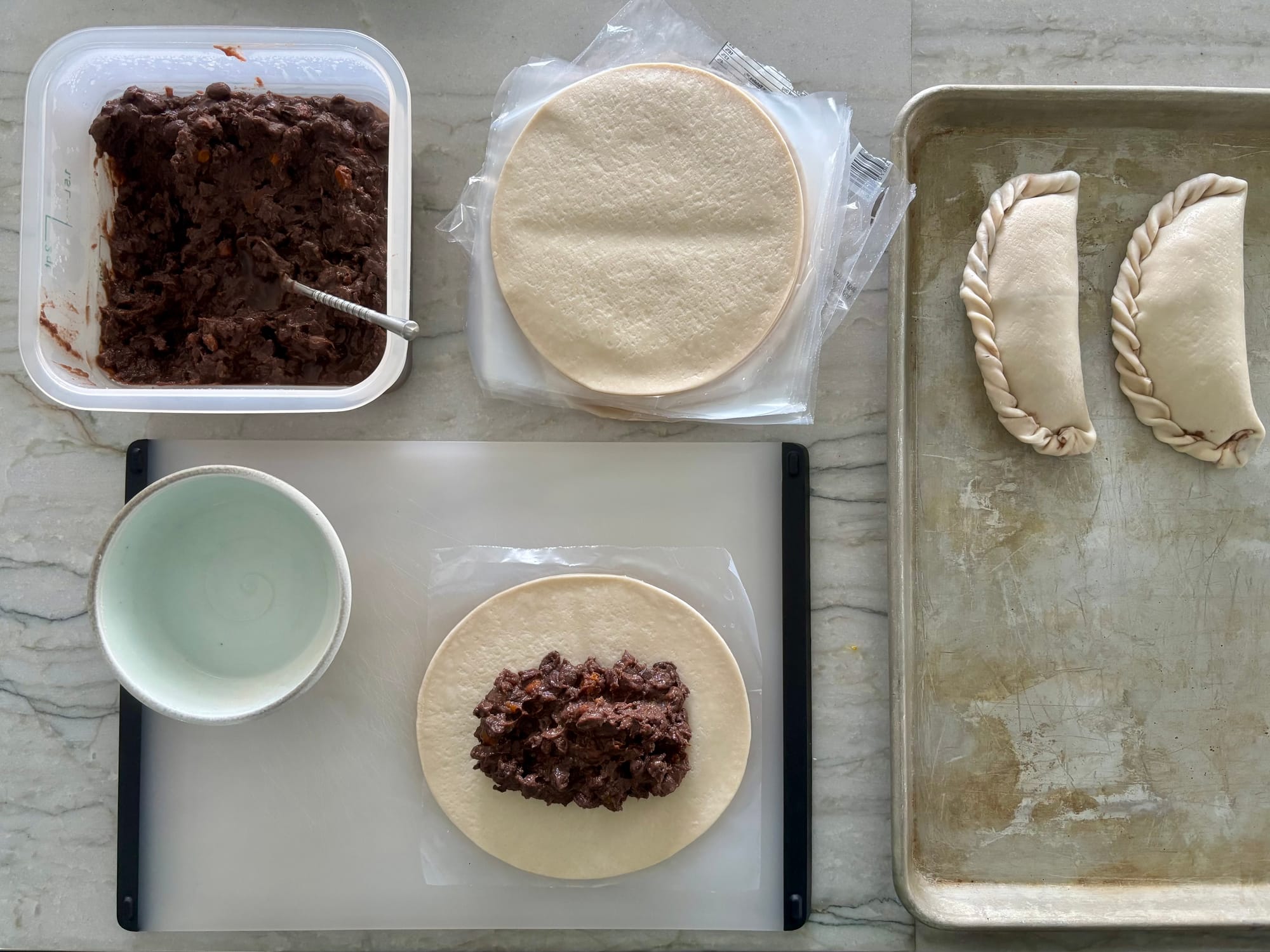 An open empanada wrapper with beans on top, sitting on a cutting board. Plus, a tub of refried beans, a stack of empanada wrappers, a bowl of water, and a sheet pan with stuffed, crimped empanadas.