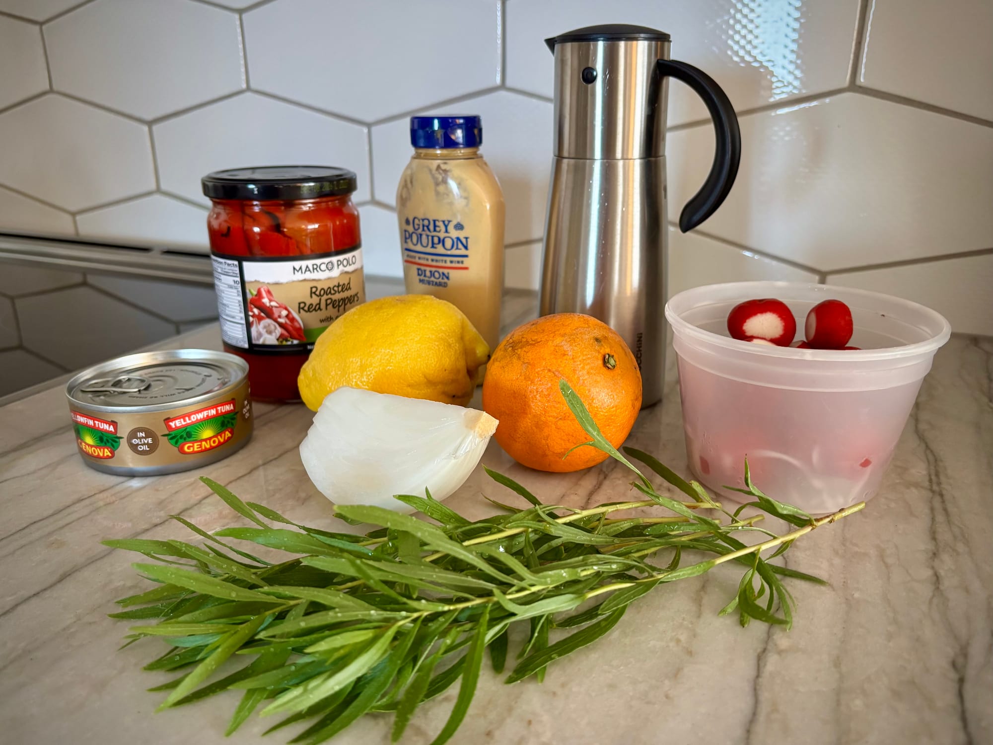 Bottles of dijon mustard and olive oil, a tub of radishes, sprigs of fresh tarragon, an orange and lemon, a hunk of white onion, a jar of roasted red peppers, and a can of tuna.