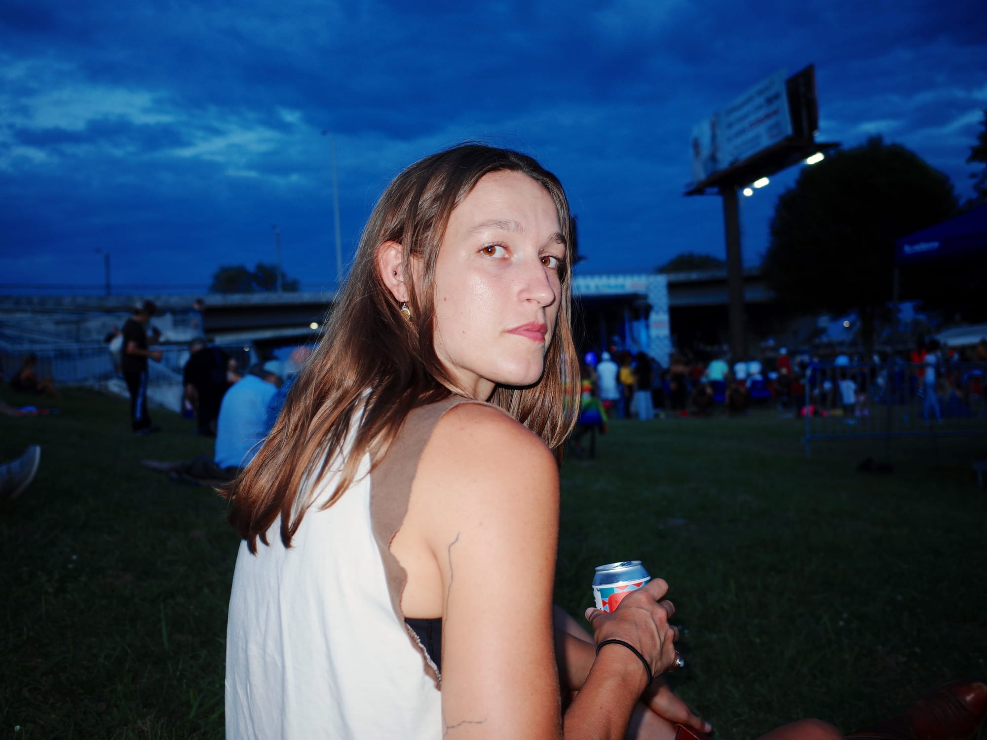 Woman (me) looking over her shoulder, sitting outside at a festival at dusk, holding a beer