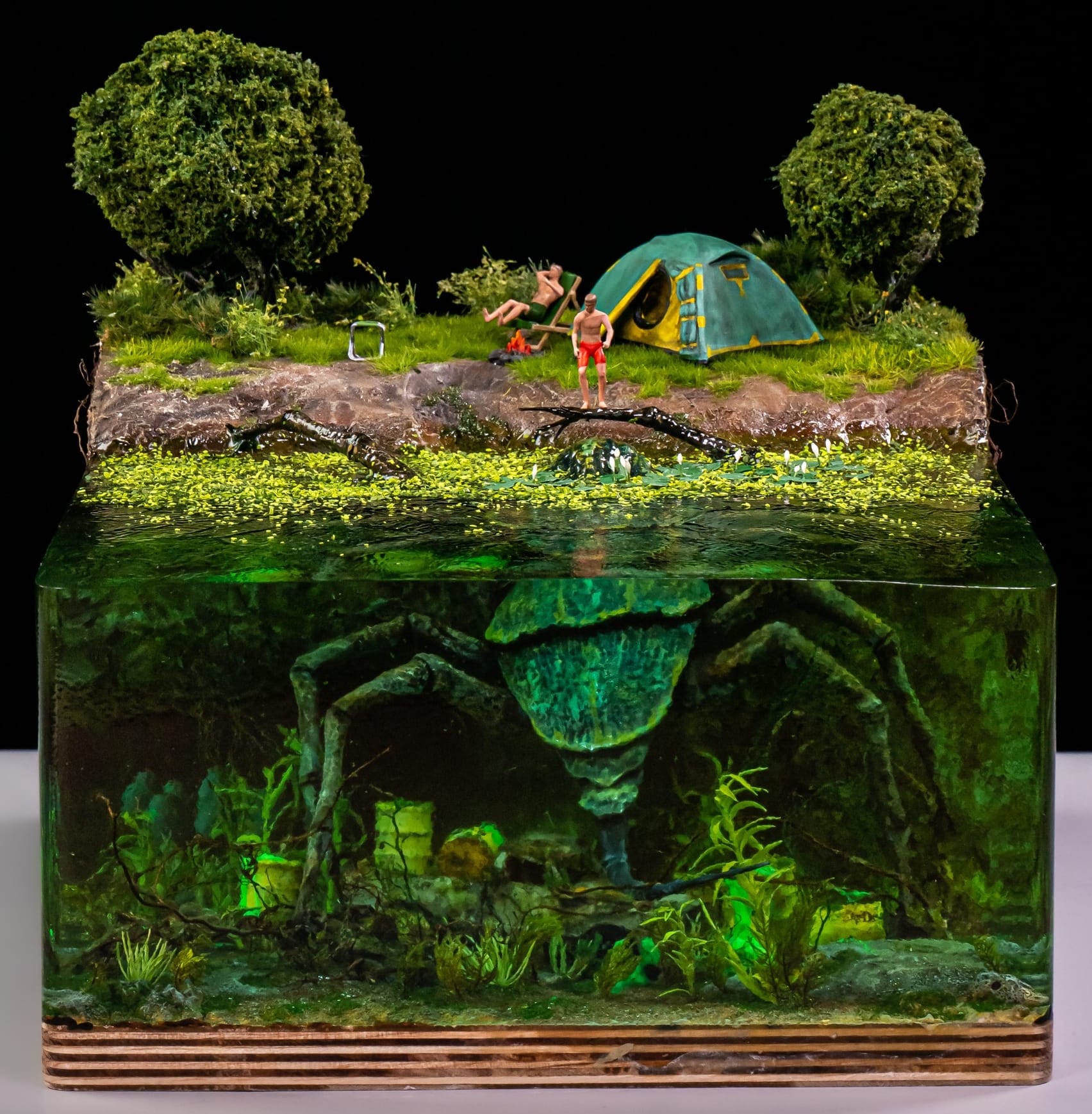 Picture of a diorama from minibricks.co, title «&nbsp;they thought it was driftwood&nbsp;». This diorama shows 2 people camping on the shore of a pond. The pond itself is made out of resin so that we can see underwater. There are barrels on the bottom of the water body and a gigantic beetle that extends its arms outside of the water in an area covered in duckweed. One of the man is standing on top of one of those arms, thinking it’s driftwood.