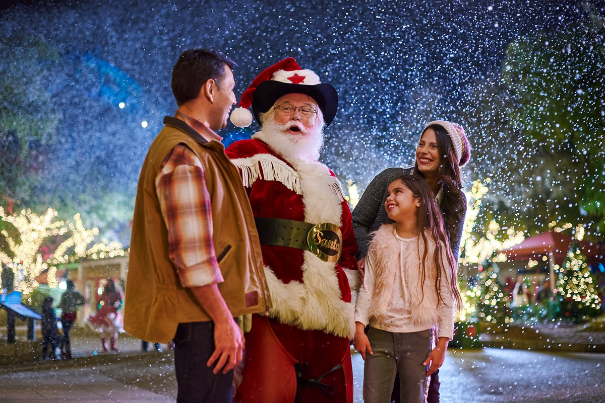 A family smiles with Santa beneath falling artificial snow and holiday lights.