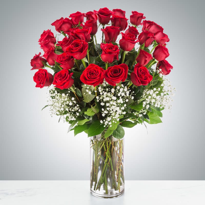A luxurious floral arrangement named "Endless Devotion," featuring three dozen long-stemmed red roses and baby's breath elegantly displayed in a tall, clear glass vase on a white marble surface.