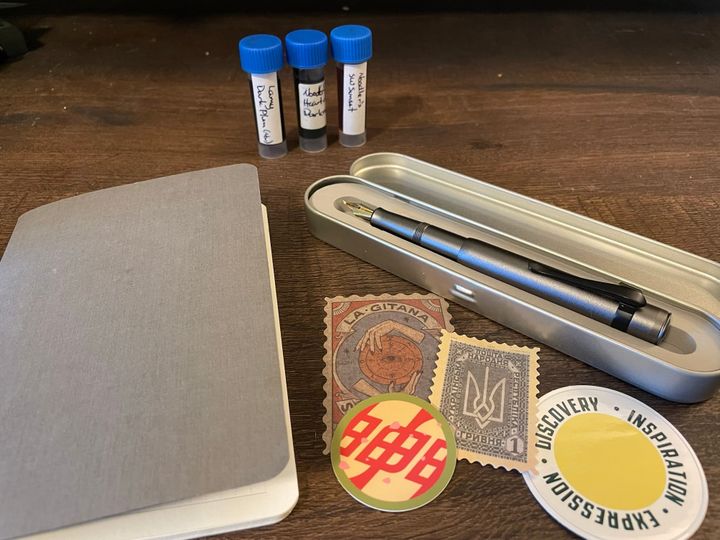 A grey notebook, stickers and a grey pen in a metal tin