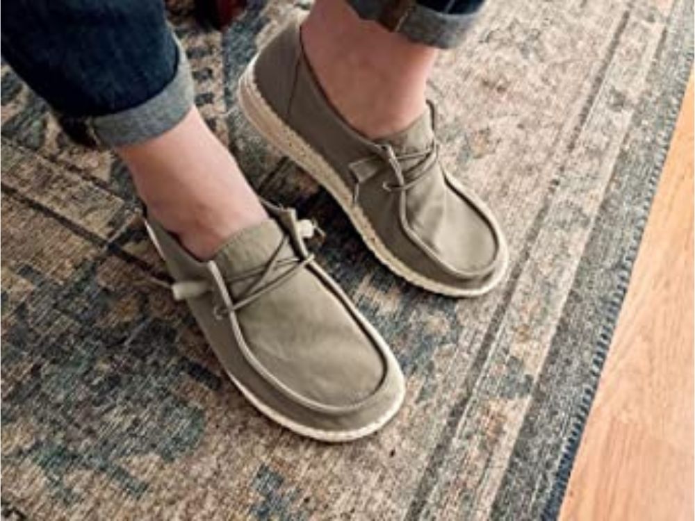 Person wearing brown Hey Dude shoes, with feet on area rug and hardwood floor.
