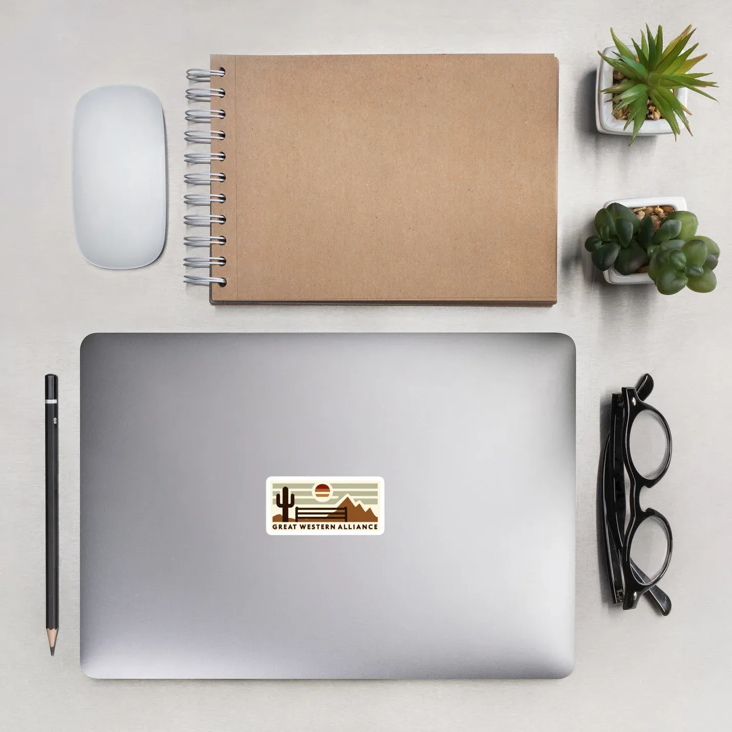 Flat lay workspace with laptop displaying Great Western Alliance logo sticker, spiral notebook, white mouse, pen, glasses, and potted succulents on white surface.