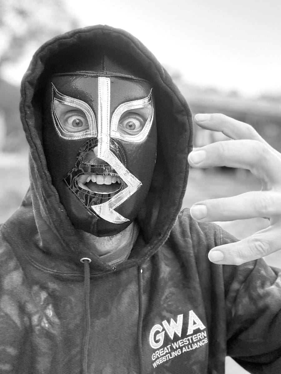 Wrestler in black and white striped luchador mask and GWWA hoodie gestures toward camera with intense expression against cloudy outdoor background.