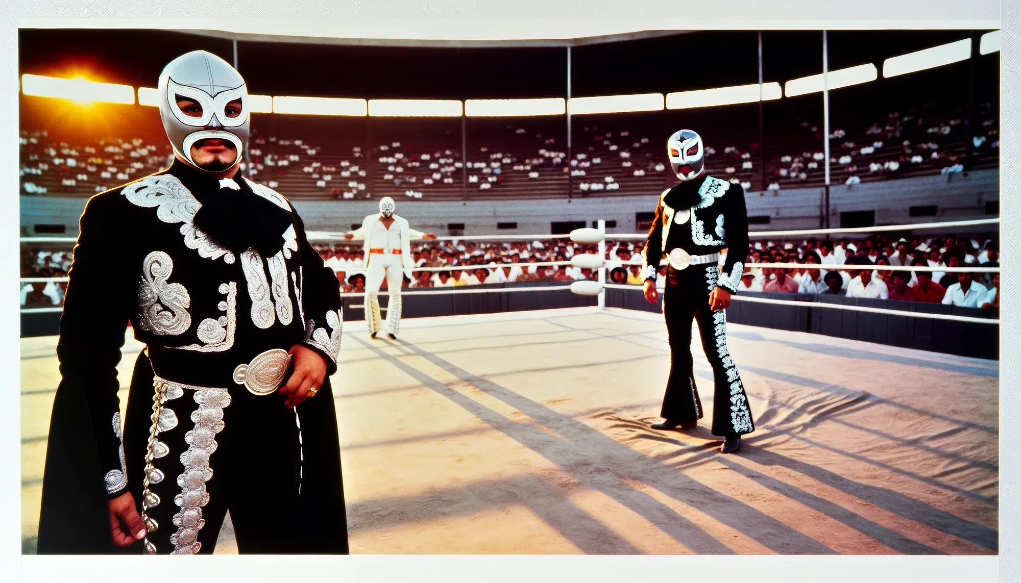 Luchador wrestlers in ornate black and silver costumes face off in a crowded arena ring at sunset.