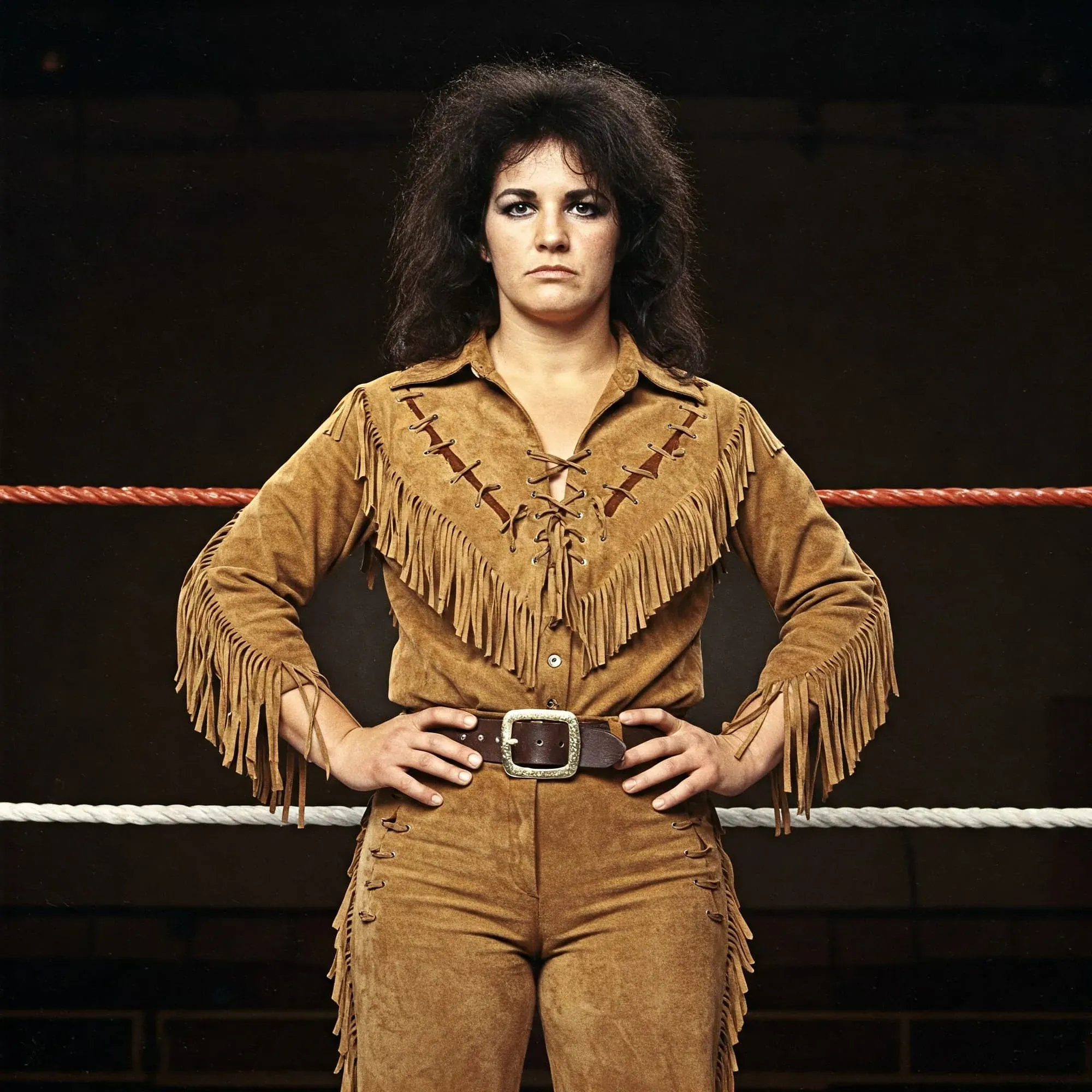 Abby Clayton stands in a wrestling ring wearing a tan suede fringe outfit and belt buckle, with dark hair and a serious expression.
