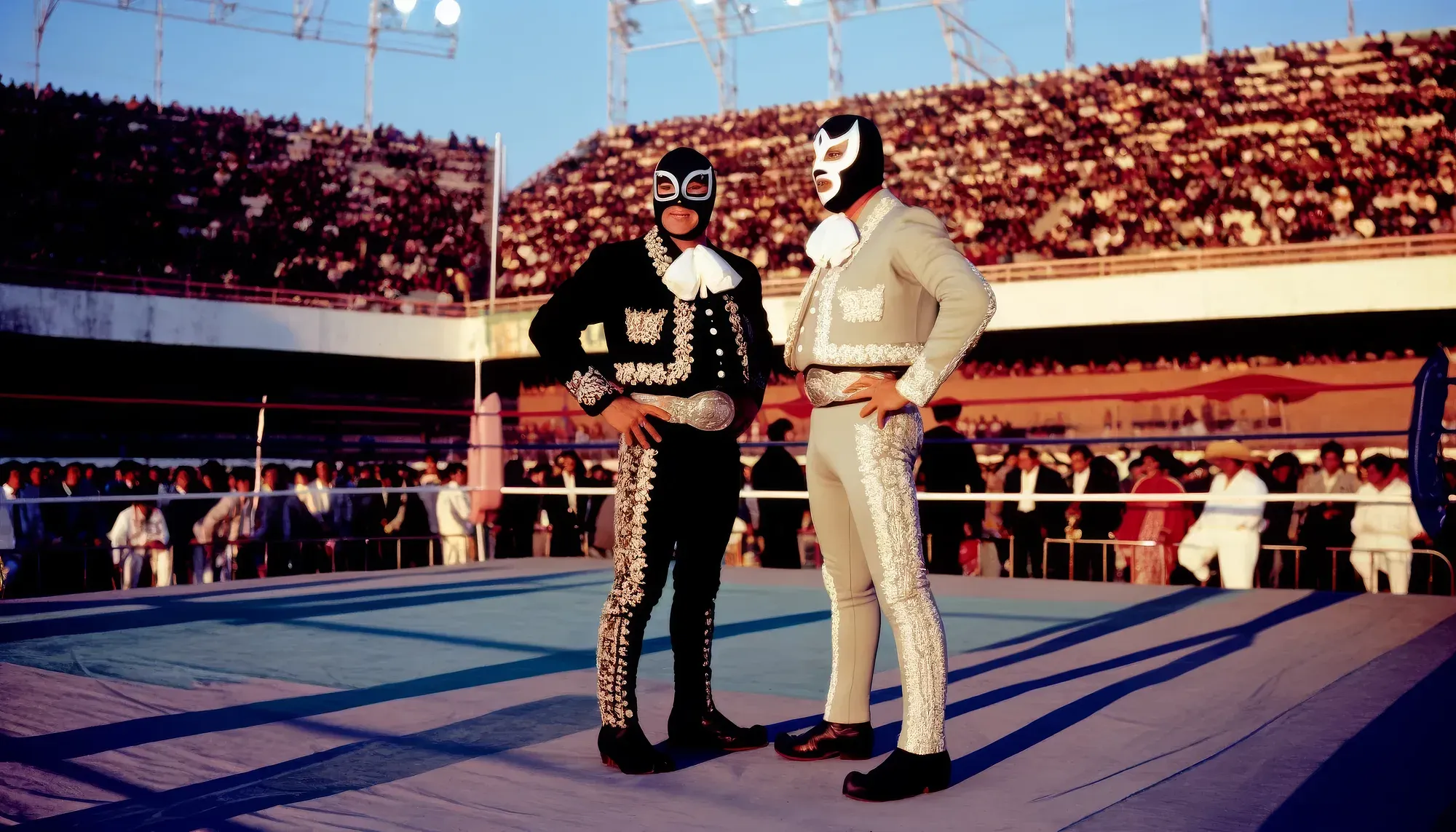 Two masked wrestlers in elaborate sequined costumes stand in a wrestling ring before a packed stadium audience.
