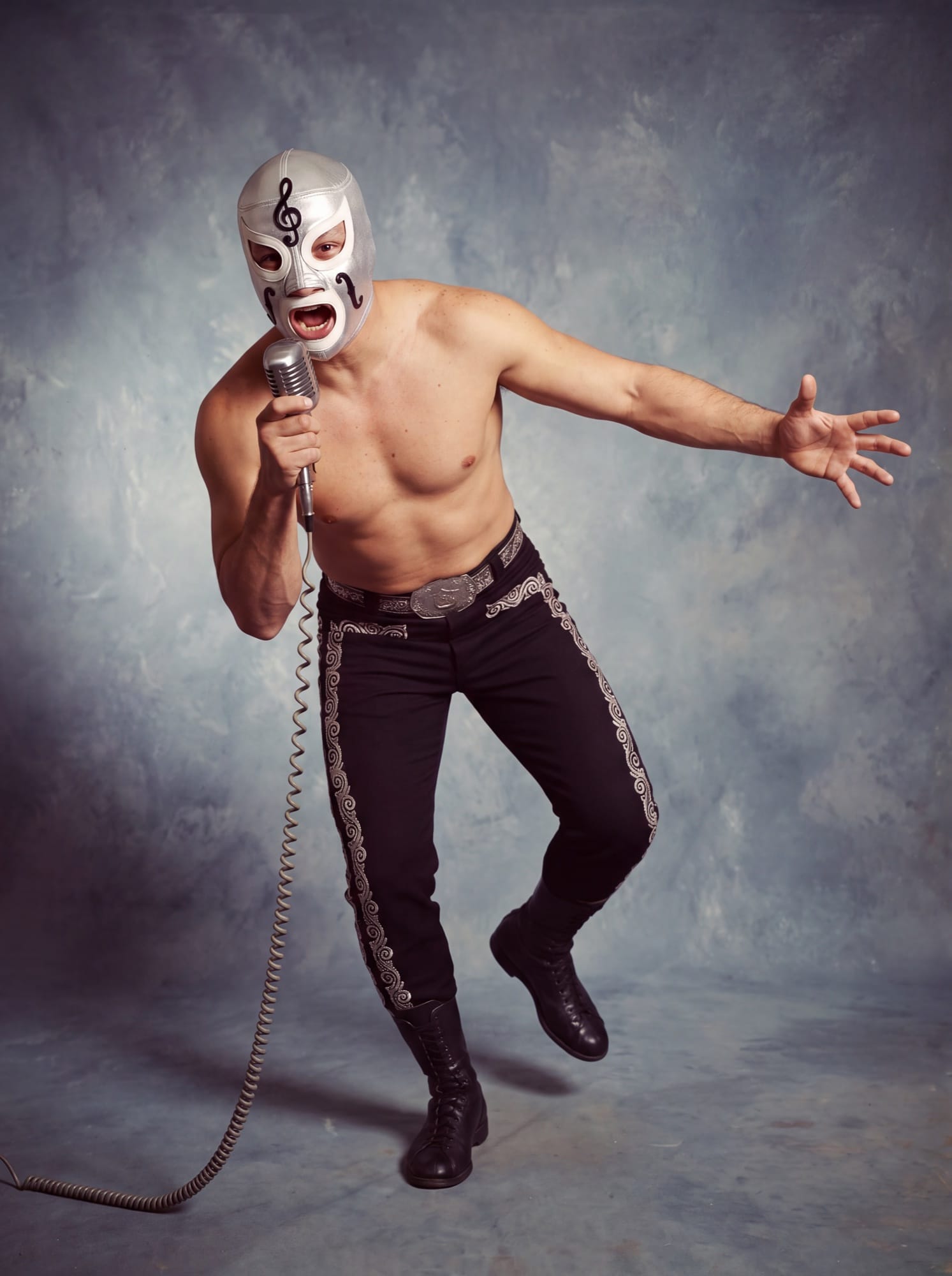El Violín in his silver treble-clef mask and black charro tights, shouting into a microphone with one arm thrown wide.