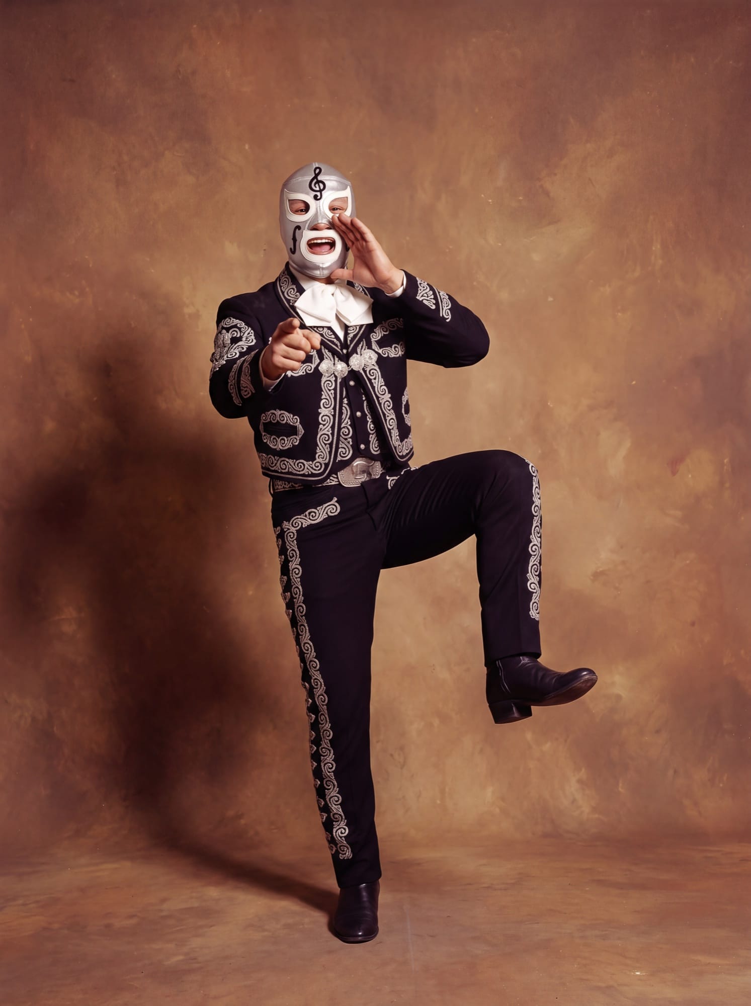El Violín in a black and silver charro suit, one knee raised, pointing at the camera with a hand cupped at his mouth.