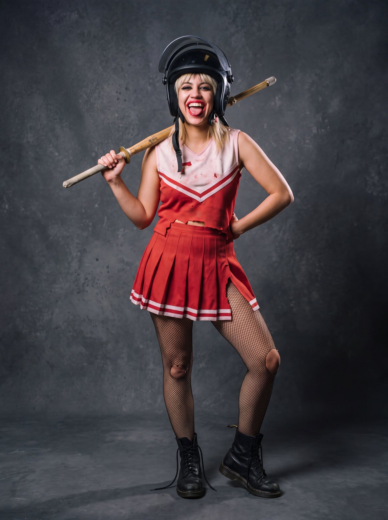 Trash Candy, a punk women's wrestler, in a black police riot helmet with the visor up, a torn red and white cheerleader uniform, ripped fishnets and combat boots, laughing at the camera with a kendo stick over her shoulder.