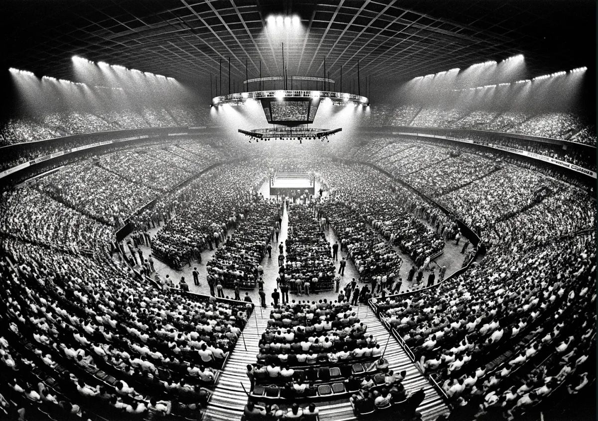 Packed arena stadium with thousands of spectators surrounding a boxing ring under bright floodlights during a 1970s wrestling event.