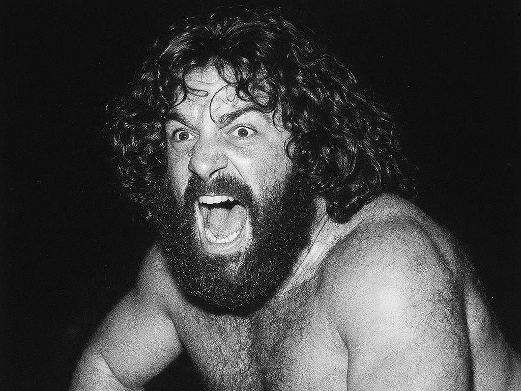 Brody Broscious, shirtless wrestler with long dark hair and full beard, makes an intense facial expression against a black background.