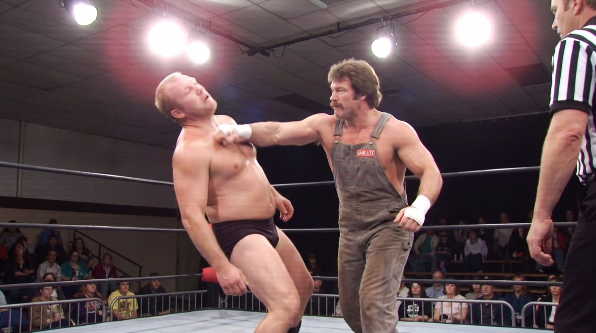 Billy Larson, in faded denim bib overalls and taped wrists, drives a forearm smash into Wendell Briggs's chest in a small 1970s arena, a referee at the edge of the ring.