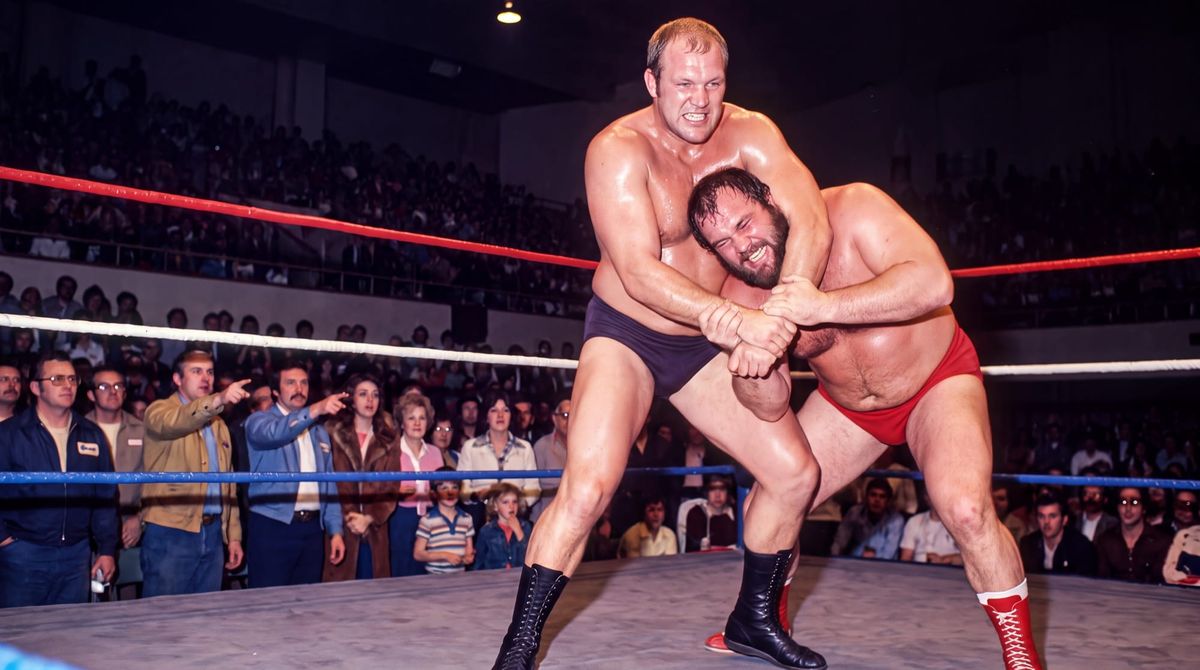 Wendell Briggs holds a bearded opponent in red trunks in a side headlock in front of a packed civic auditorium crowd, 1978.
