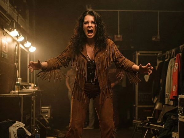 Abby Clayton stands in a dressing room wearing a brown fringe jacket and studded top, arms spread wide with an excited expression.