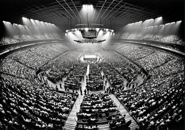 Packed arena stadium with thousands of spectators surrounding a boxing ring under bright floodlights during a 1970s wrestling event.
