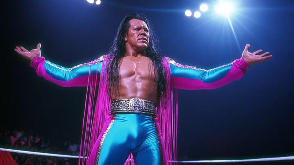Jesse Stardust stands in a wrestling ring with arms spread wide, wearing a magenta and turquoise fringed robe with a decorative belt.
