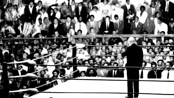 Bald wrestler in dark suit stands in wrestling ring facing crowd in packed stadium during 1970s event.
