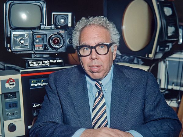 Franklin J. Lewis sits in a television studio surrounded by vintage broadcast equipment, wearing a dark suit and striped tie.