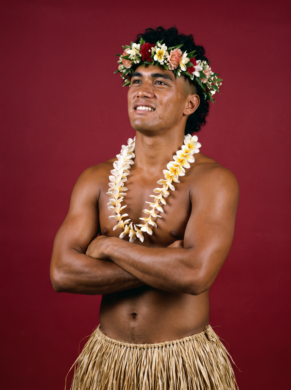 Kalani Koa in a haku lei flower crown, plumeria lei, and grass skirt, arms crossed against a deep red studio backdrop in a 1989 promotional photograph