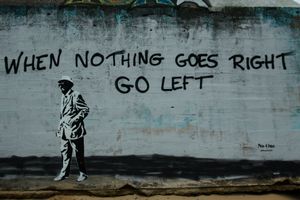 A wall painted with the words When Nothing Goes Right Go Left. A old man in a hat is also painted on the wall. He is walking to the left.