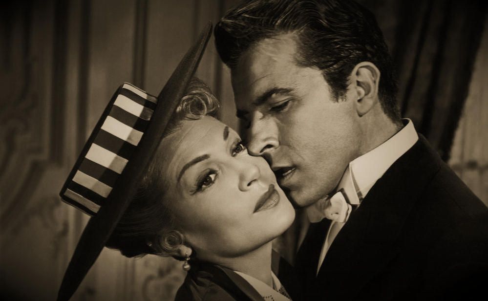 Fernando Lamas de smoking, mejilla contra mejilla con una actriz rubia de sombrero, en una escena de película