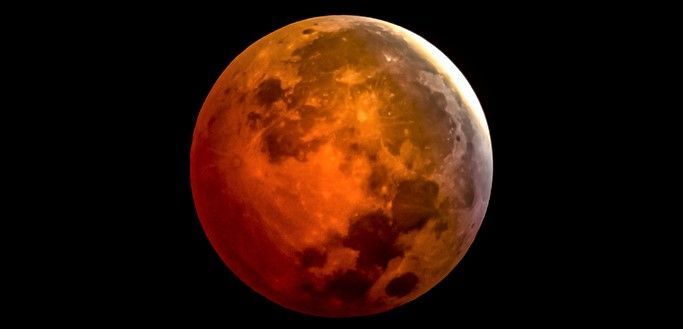 Luna llena teñida de naranja rojizo durante un eclipse total, con el borde superior más claro, sobre un cielo completamente negro.