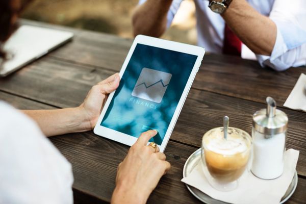Una mujer consulta un gráfico financiero en una tablet sobre una mesa de madera, con un café al lado y otra persona sentada enfrente.