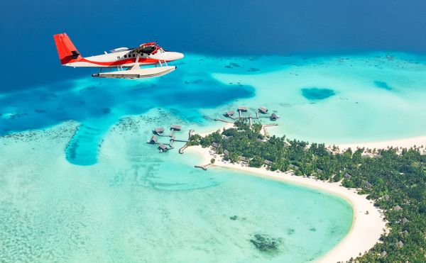 Un hidroavión rojo y blanco sobrevuela un atolón de Maldivas, con bungalows sobre el agua turquesa y una playa de arena blanca.
