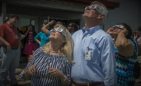 Un grupo de personas mira hacia el cielo con gafas de eclipse de montura blanca, al aire libre y a plena luz del día.