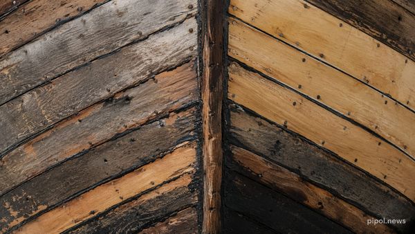 Detalle del casco de un barco de madera donde conviven tablas grises y envejecidas con otras claras y recién colocadas, unidas por juntas de calafateo negro.
