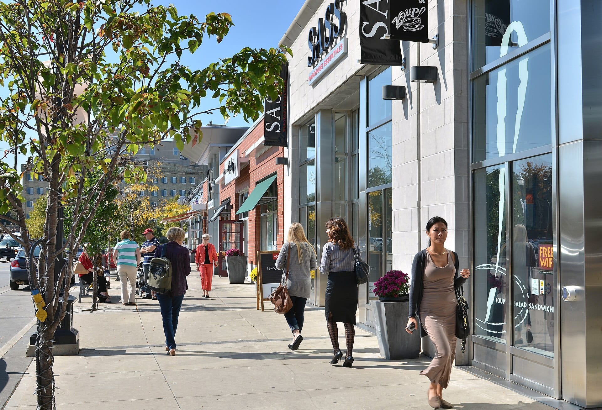 Shoppers walking along a sunny retail street, passing several storefronts with only some stepping inside.
