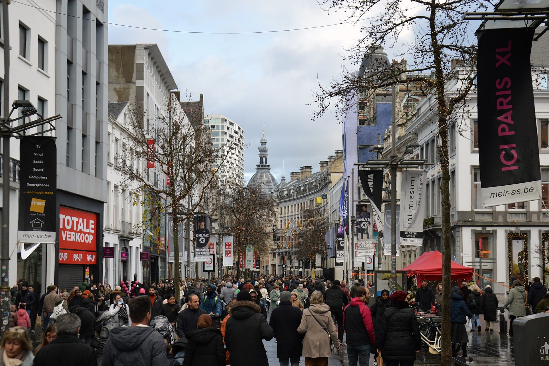A dense crowd of holiday shoppers walking down a pedestrian shopping street lined with stores and banners.