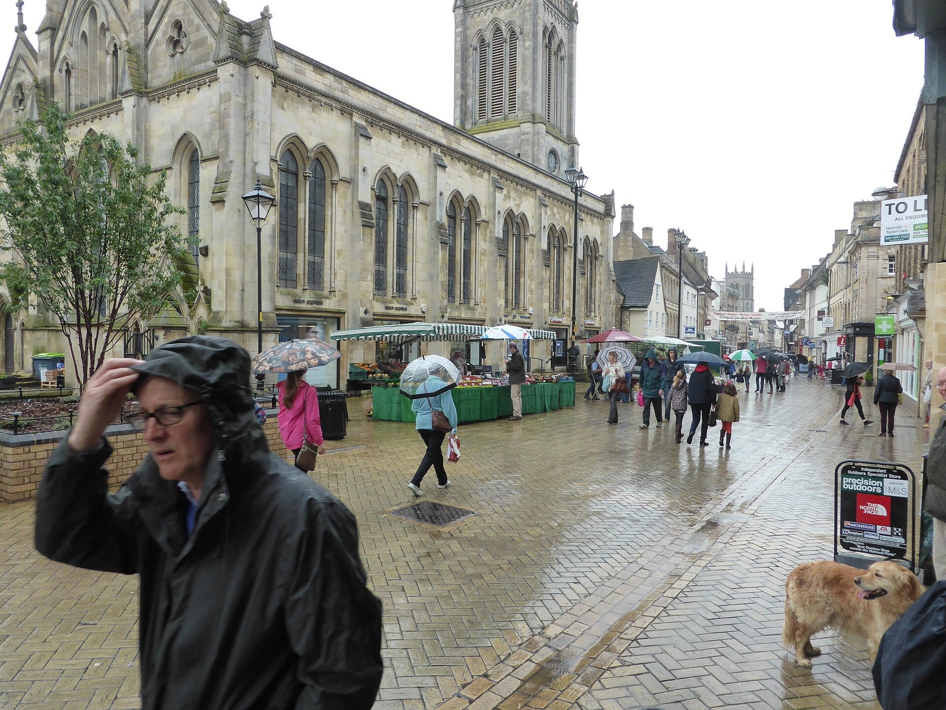 Shoppers with umbrellas and rain jackets walking along a wet brick pedestrian shopping street on a rainy day.