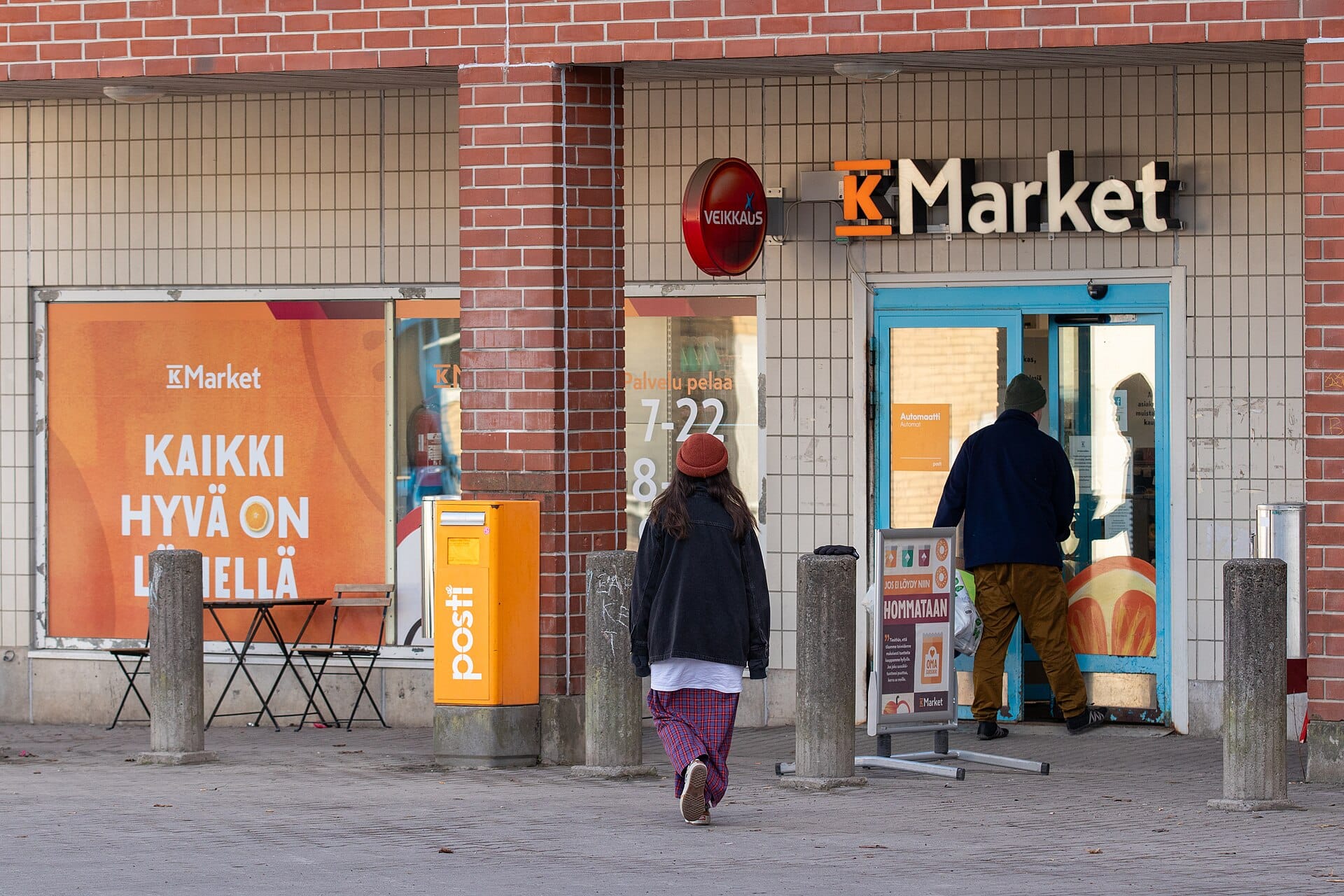 Two shoppers walking into a grocery store entrance from a brick-paved sidewalk