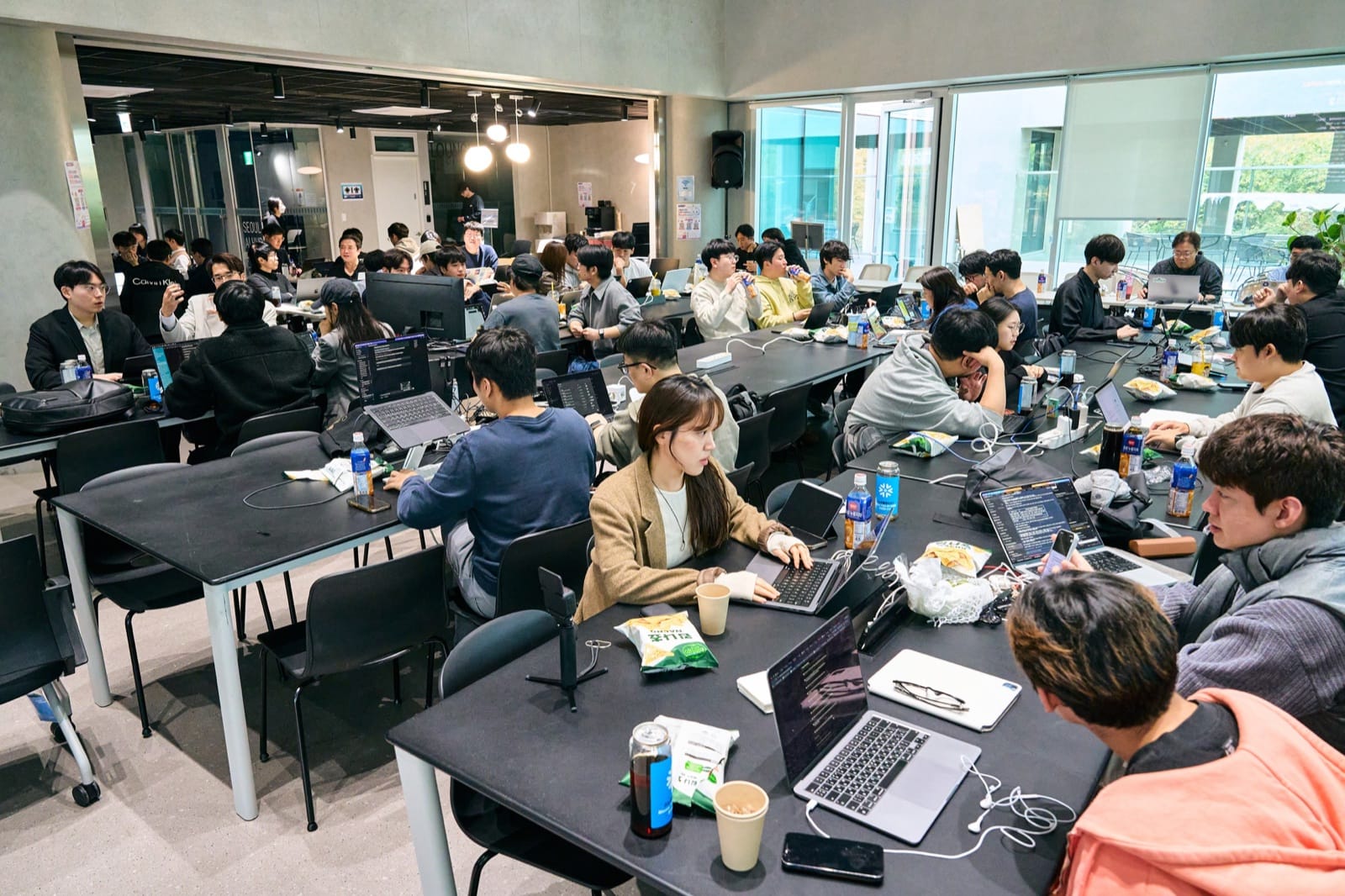 A room packed with participants building at long tables, laptops open and snacks and drinks between them