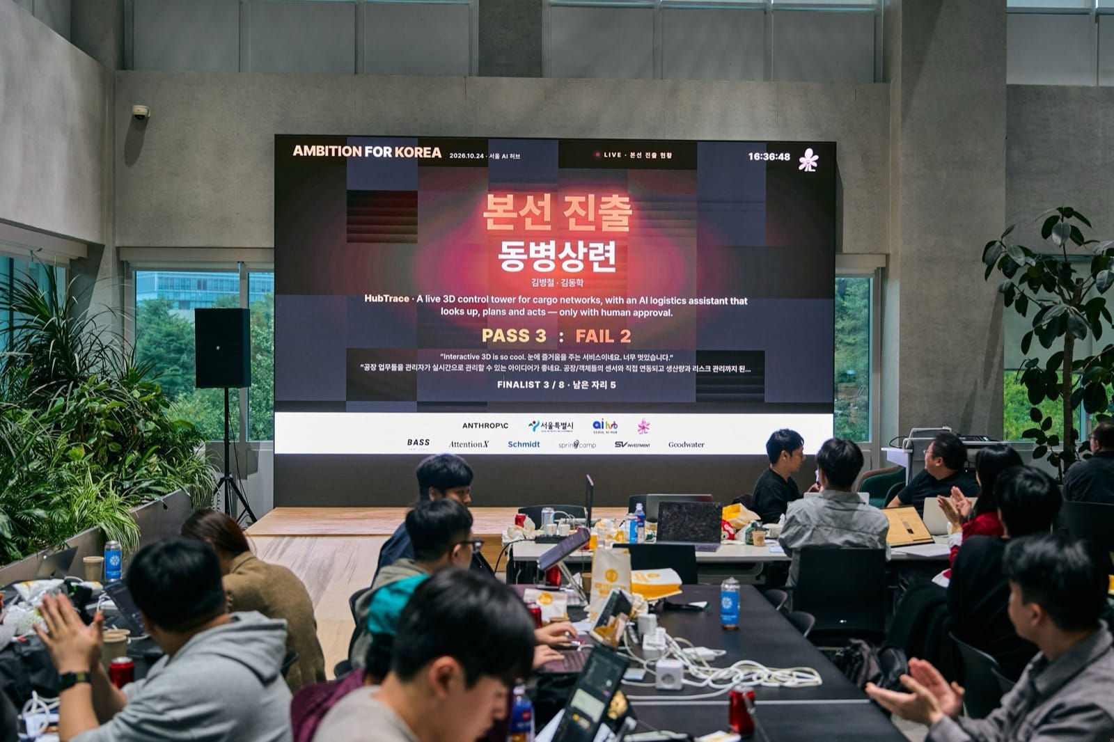 Participants at laptops under an LED wall announcing Dongbyeongsangryeon as a finalist with 3 PASS and 2 FAIL