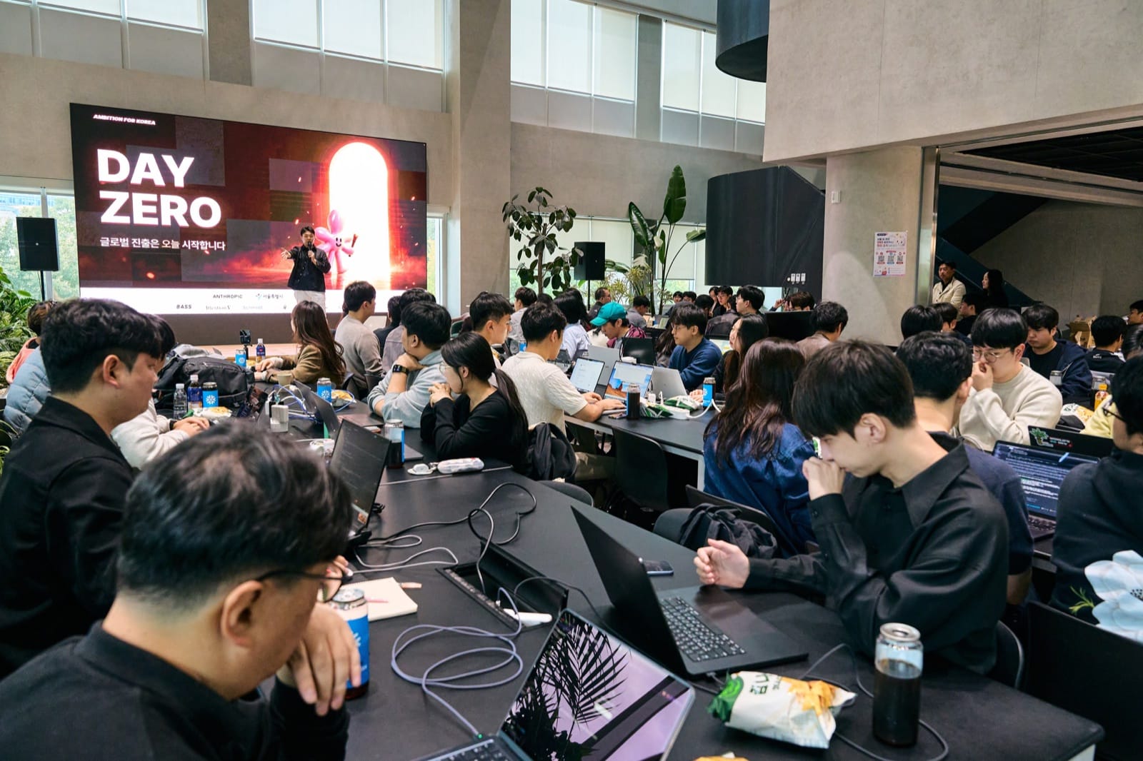 Participants at long tables with laptops open, listening to the opening under an LED wall that reads DAY ZERO