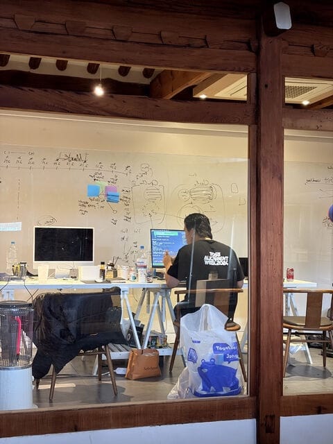 Seen through a glass wall, someone working on a laptop at a desk in front of a whiteboard covered in notes
