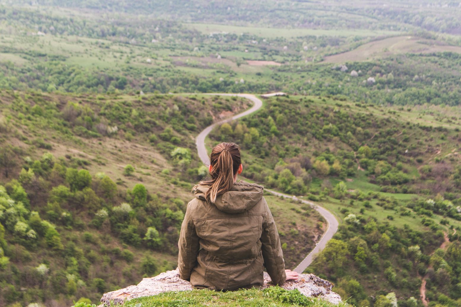 A girl sitting on a rock overlooking a valley with a winding road