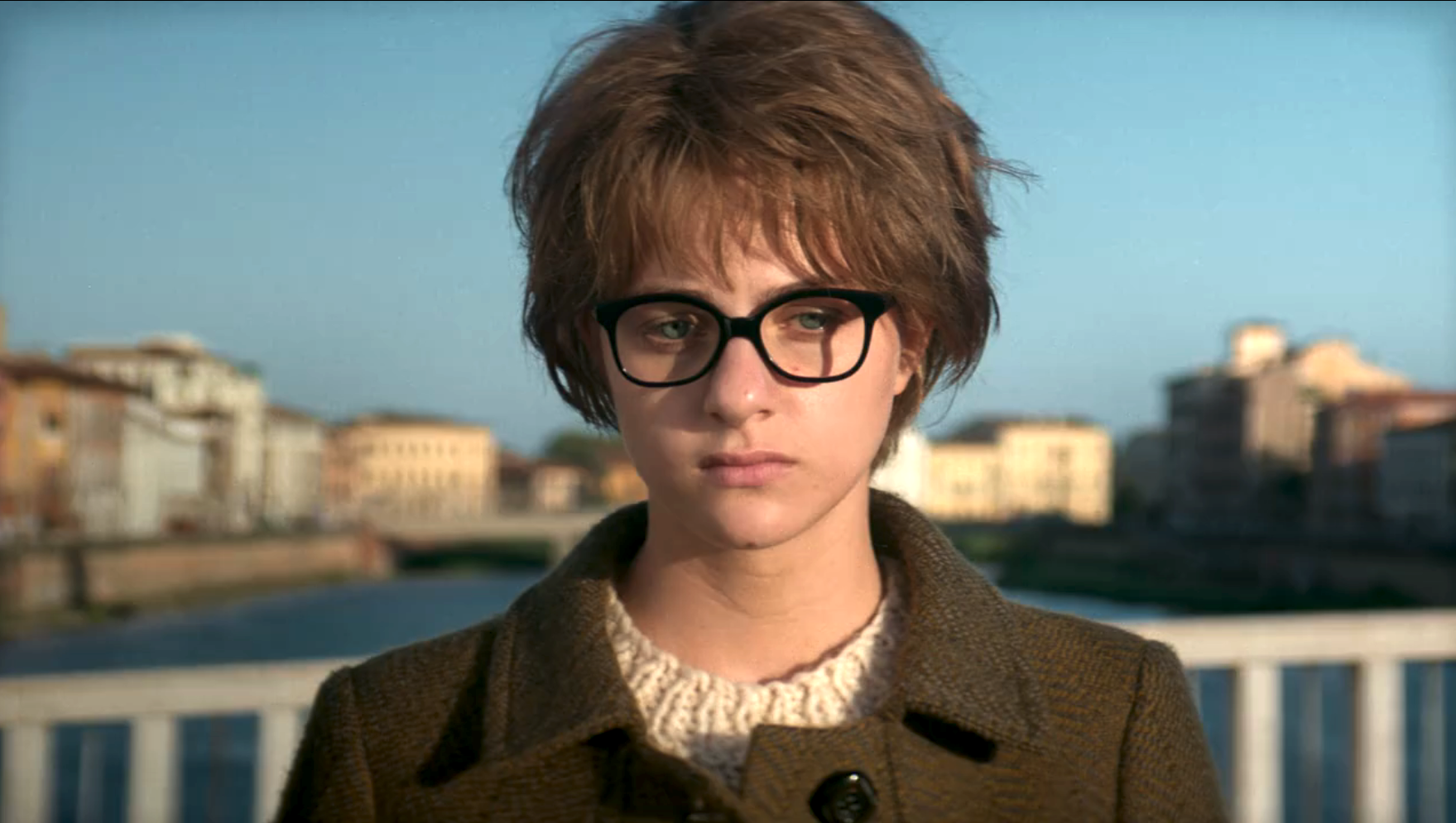 Lenu in medium close up standing on the Solferino Bridge overlooking the Arno, wearing a sweater, wool coat, black glasses, and a janky short wig.