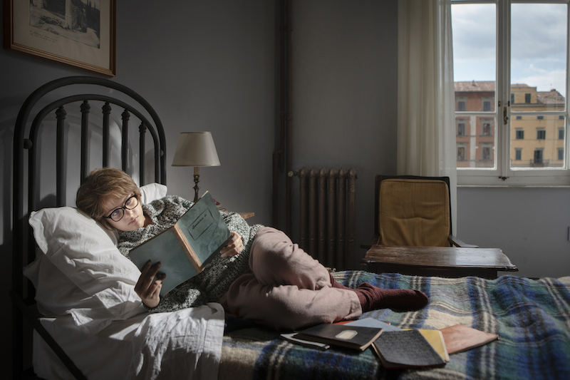 Lenu reads Lila's notebooks in bed during the day on top of a blue plaid wool blanket.