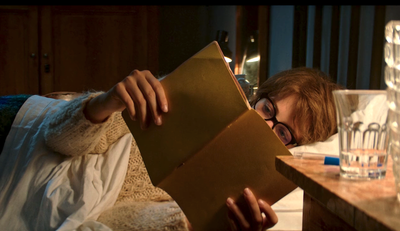 Lenu reads Lila's notebooks in bed at night while wearing a white wool sweater.